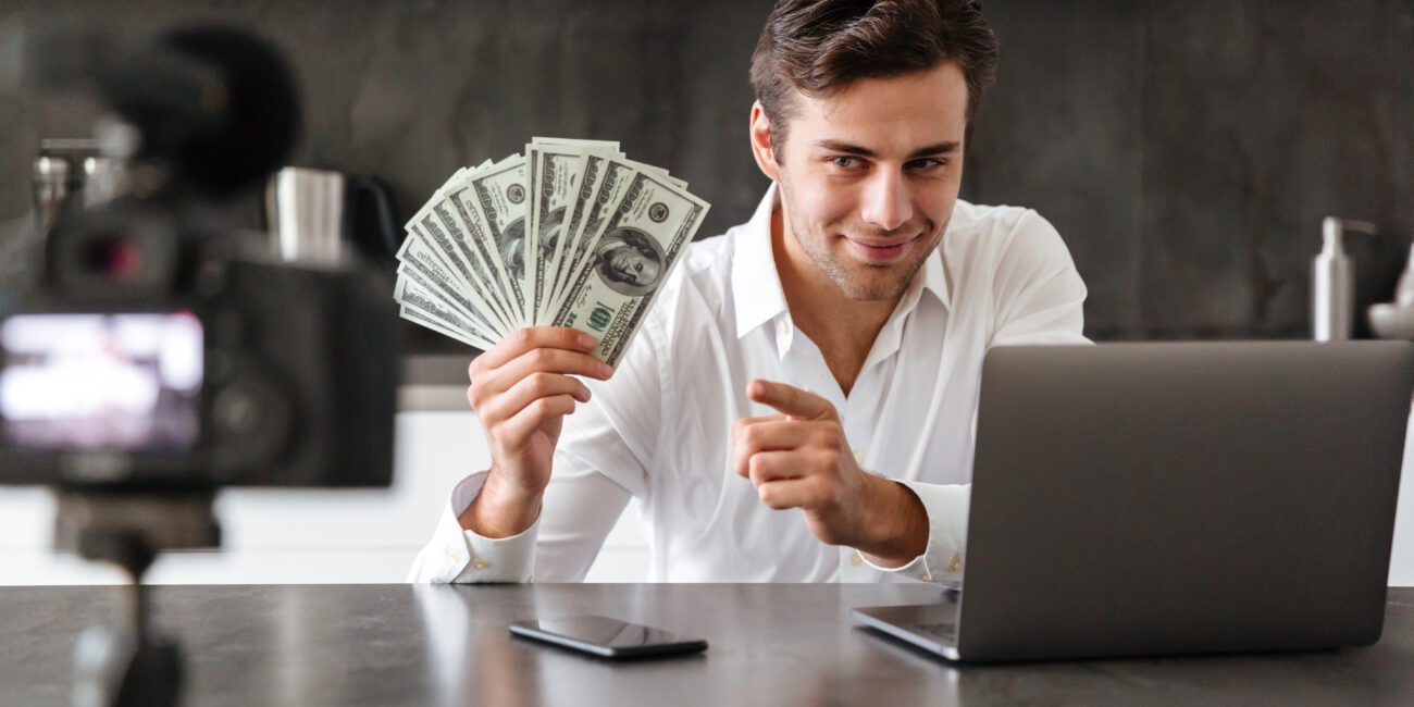 Smiling young man filming his video blog episode about new tech devices while sitting at the kitchen table with laptop and showing bunch of money banknotes
