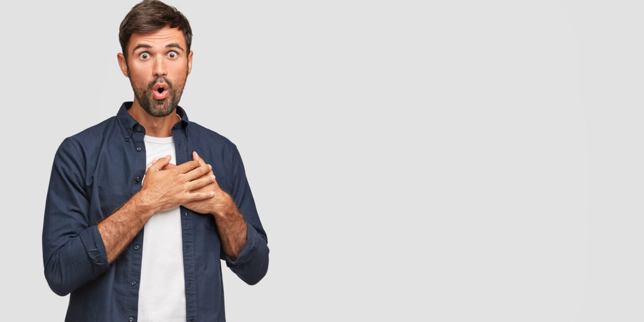 Stunned emotive unshaven man with shocked expression, keeps both palms on heart, sees something awesome in front, isolated over white background with copy space for your promotional content.