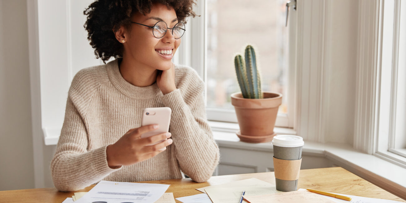 People, ethinicity and entertainment concept. Beautiful delighted thoughtful pleased female financier checks updates for organiser on mobile phone, sits at desk with documents, studies graphics